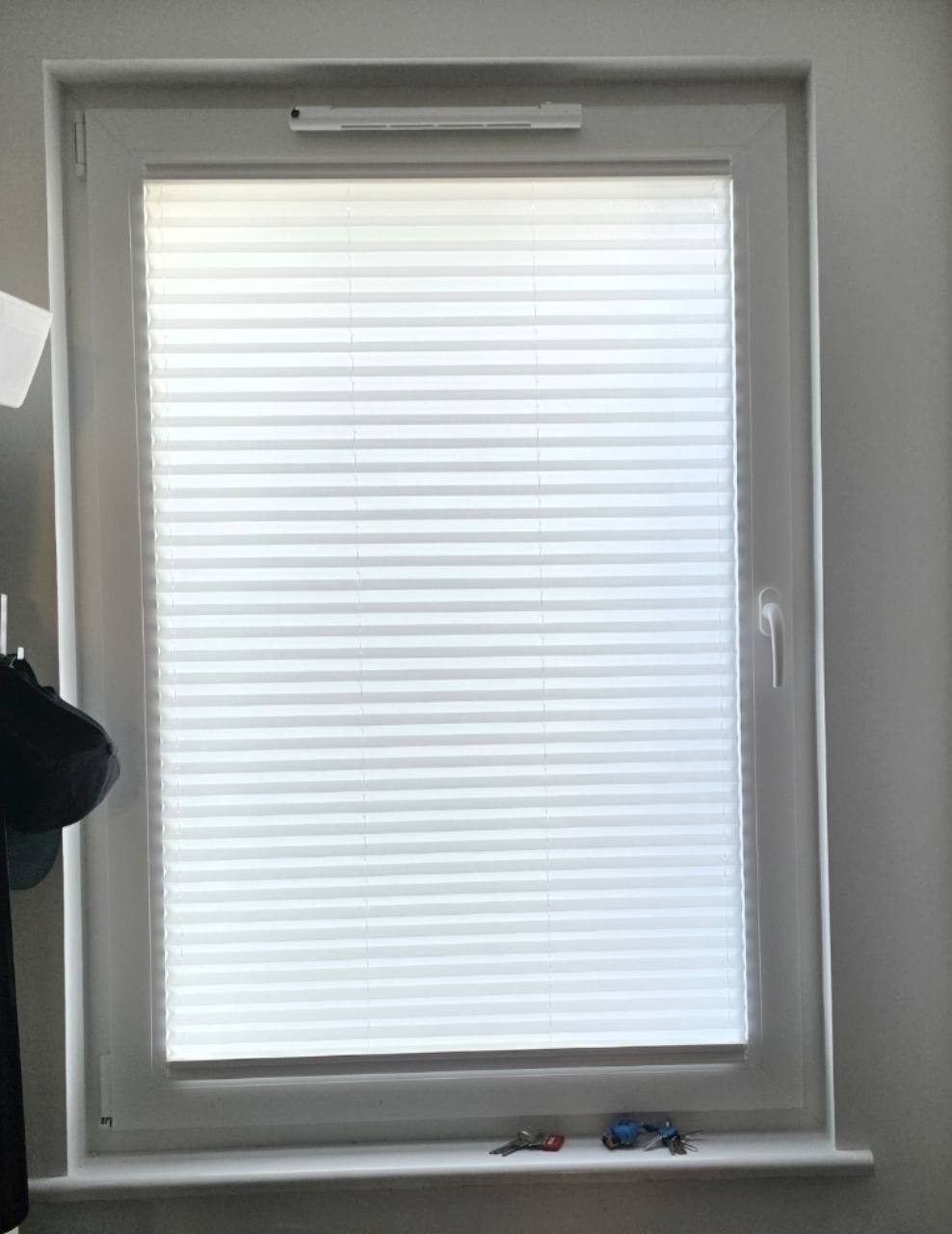 Biała plisa zasłaniająca szybę pionowego okna w białej ramie.